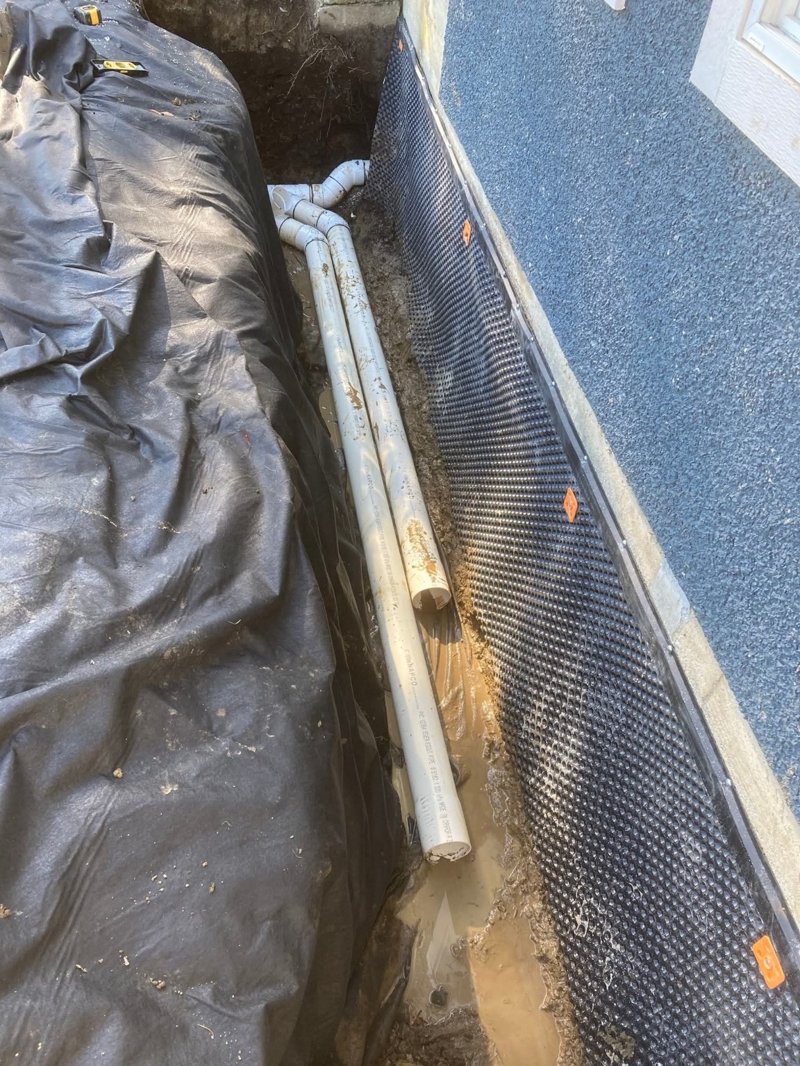Dimpled foundation drainage membrane beside white PVC pipes in a perimeter trench