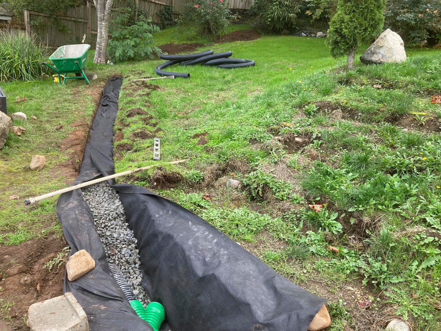 French drain installation with geotextile, aggregate, black pipe and a green pipe junction